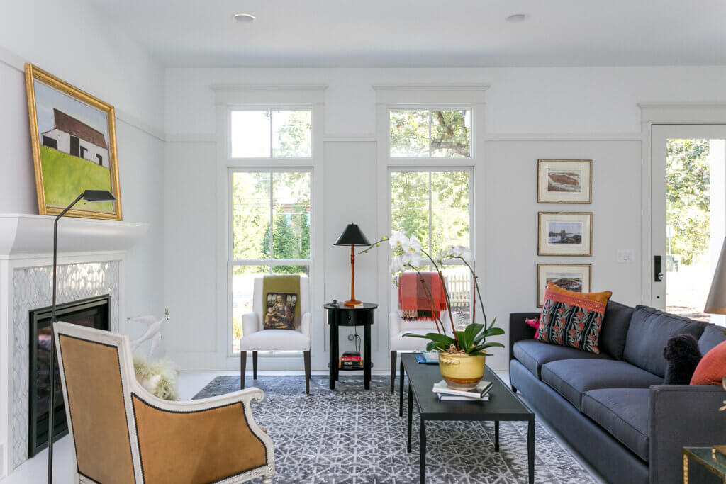 A brightly lit living room with large windows, white walls, and a grey patterned rug. It features a grey sofa adorned with colorful pillows, a glass coffee table with a potted plant, two white chairs, a fireplace with a painting above it, and framed artwork on the walls. By Zinn Design Build.