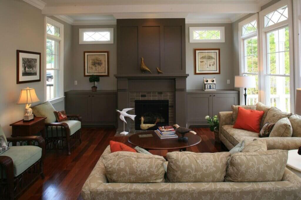 A cozy living room with a fireplace centered between two windows. The room features a neutral color palette with beige patterned sofas, a wooden coffee table, various decorative items, and framed artwork. Two armchairs sit on either side of the fireplace. Designed by Zinn Design Build