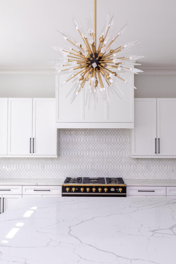 A modern kitchen featuring white cabinetry with black handles, a marble countertop, and a gold-accented stove. The backsplash has a white geometric pattern, and a starburst chandelier with gold and clear elements hangs from the ceiling. By Zinn Design Build.