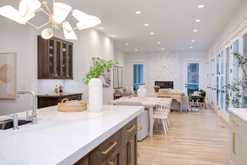 A modern open-concept kitchen and living area with white walls and wood flooring. The kitchen features a large island with white countertops, dark wooden cabinets, and contemporary light fixtures. The living area has a fireplace, light-colored furniture, and large windows. By Zinn Design Build.