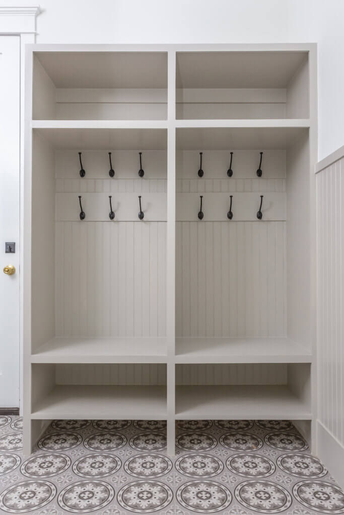 A white mudroom with two open cubbies, each featuring upper and lower shelves, and black hooks lining the middle section for hanging items. The floor is patterned with a gray and white tile design by Zinn Design Build