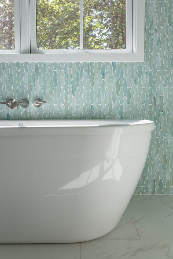 A modern bathroom featuring a white freestanding bathtub placed in front of a tiled wall with elongated hexagon-shaped tiles in shades of green and blue. A window above the bath allows natural light to illuminate the space, highlighting the sleek design by Zinn Design Build.