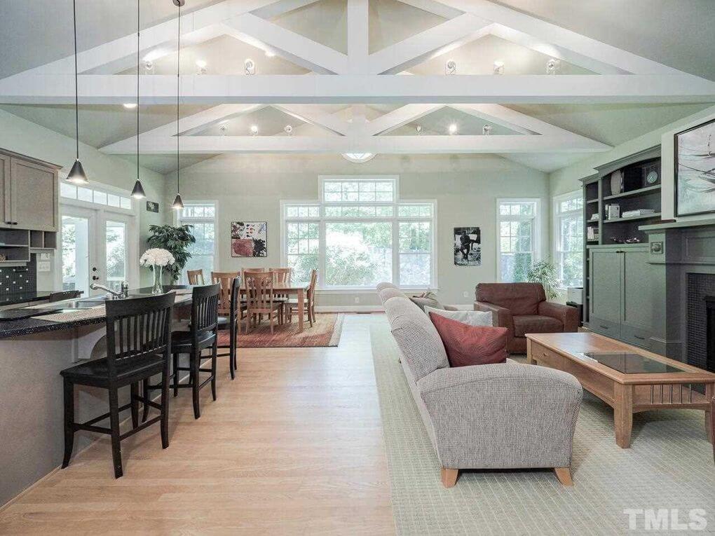 Spacious living room with vaulted ceiling and exposed beams, featuring a kitchen counter with barstools on the left, a dining table near large windows, a cozy sitting area with a sofa and armchairs, and a fireplace with built-in shelves on the right.