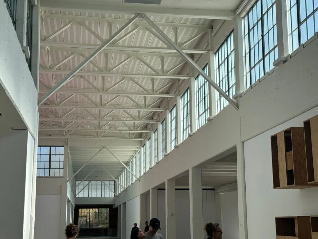 A modern interior with high ceilings and large windows, allowing natural light to illuminate the space. Exposed beams add to the industrial feel. People are scattered throughout, observing the surroundings.