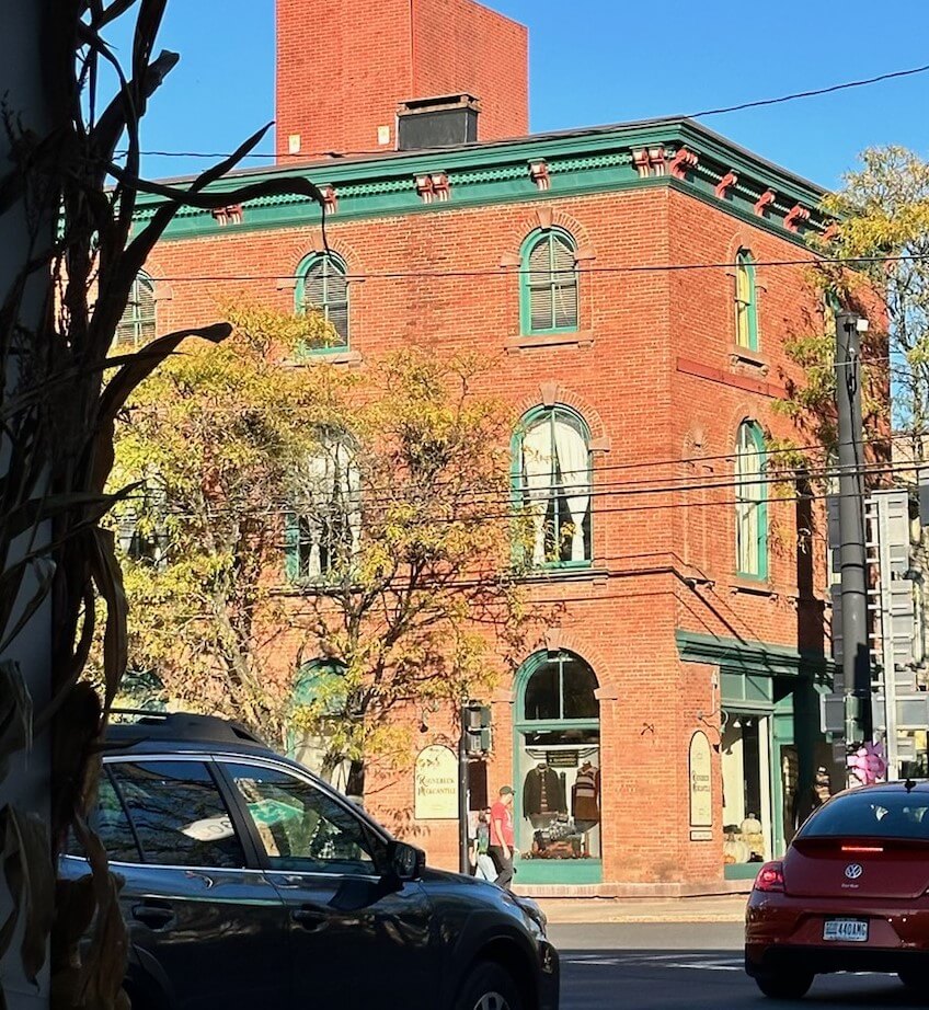 A red brick building with green window frames is seen across a street. Cars are parked in front, and a few pedestrians walk by. Leafless trees are nearby, and the sky is clear and blue, indicating a sunny day.