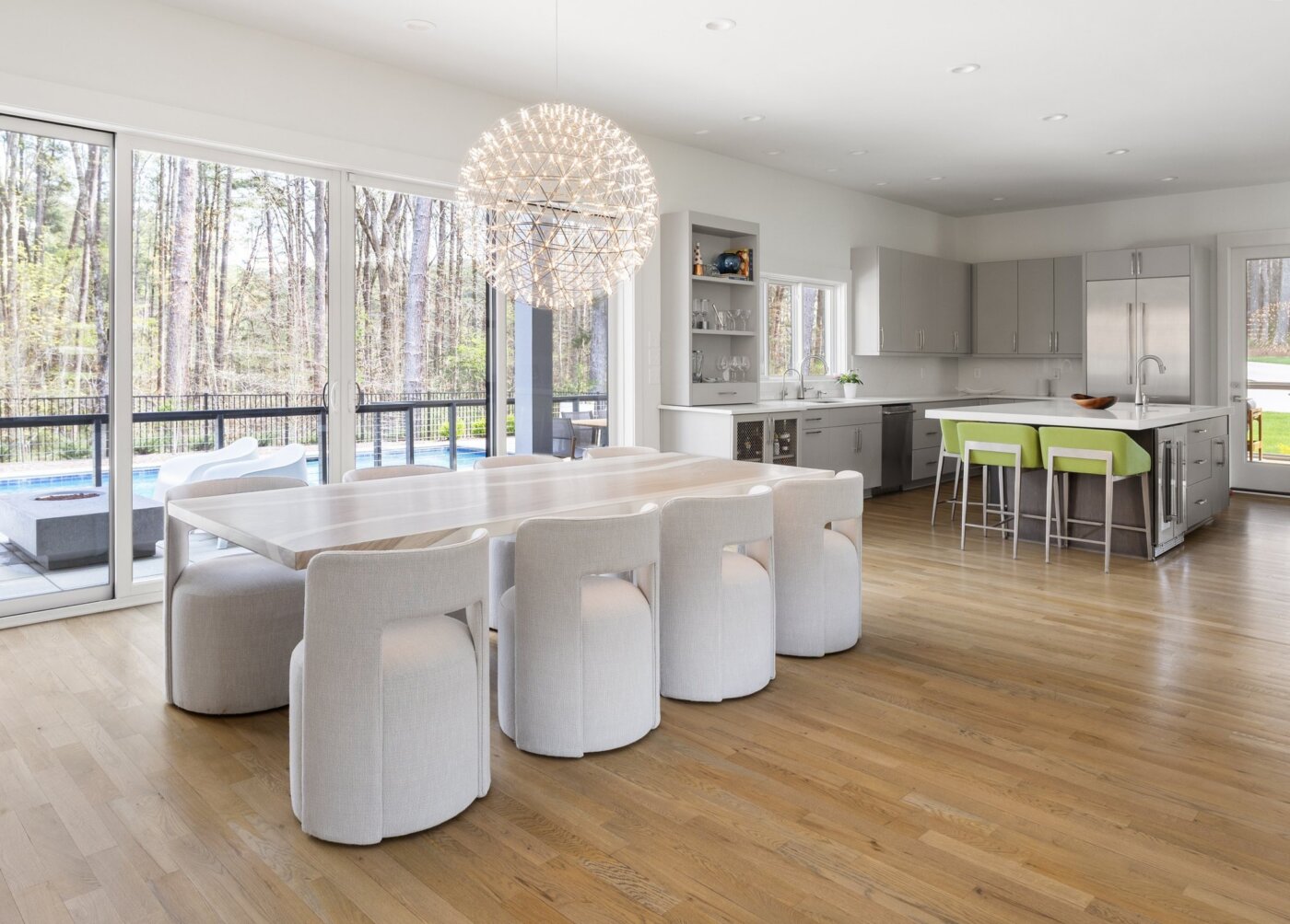 Bright modern kitchen and dining area with light wood floors, large windows, white cabinetry, a long dining table with eight white chairs, and an island with green stools. A pool and trees are visible outside.