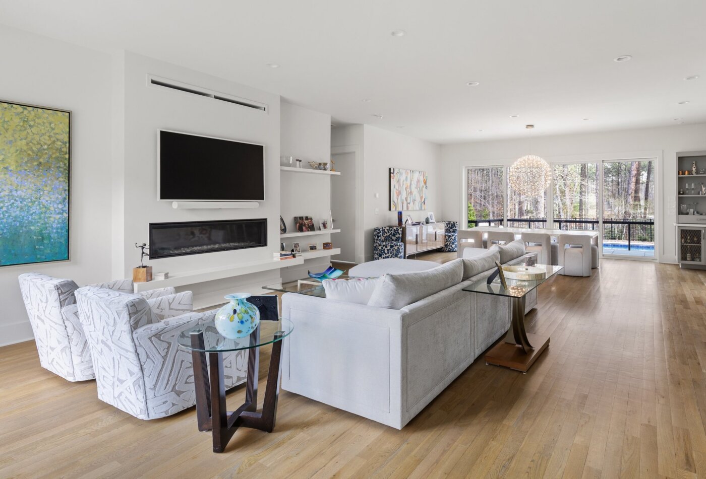 Modern living room with light wood floors, gray sofas, patterned armchairs, a wall-mounted TV above a fireplace, large windows, and contemporary art. A dining area with a chandelier is visible in the background.