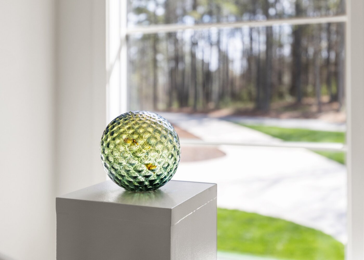A textured green glass sphere sits on a white pedestal near a window, with a blurred view of trees, grass, and pavement outside.