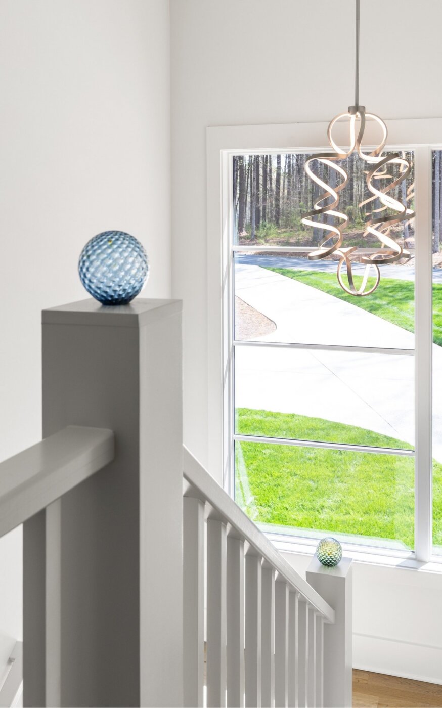 A modern staircase with white railings, a blue glass orb on the newel post, a large window with a view of green grass, and a sculptural pendant light hanging from the ceiling.