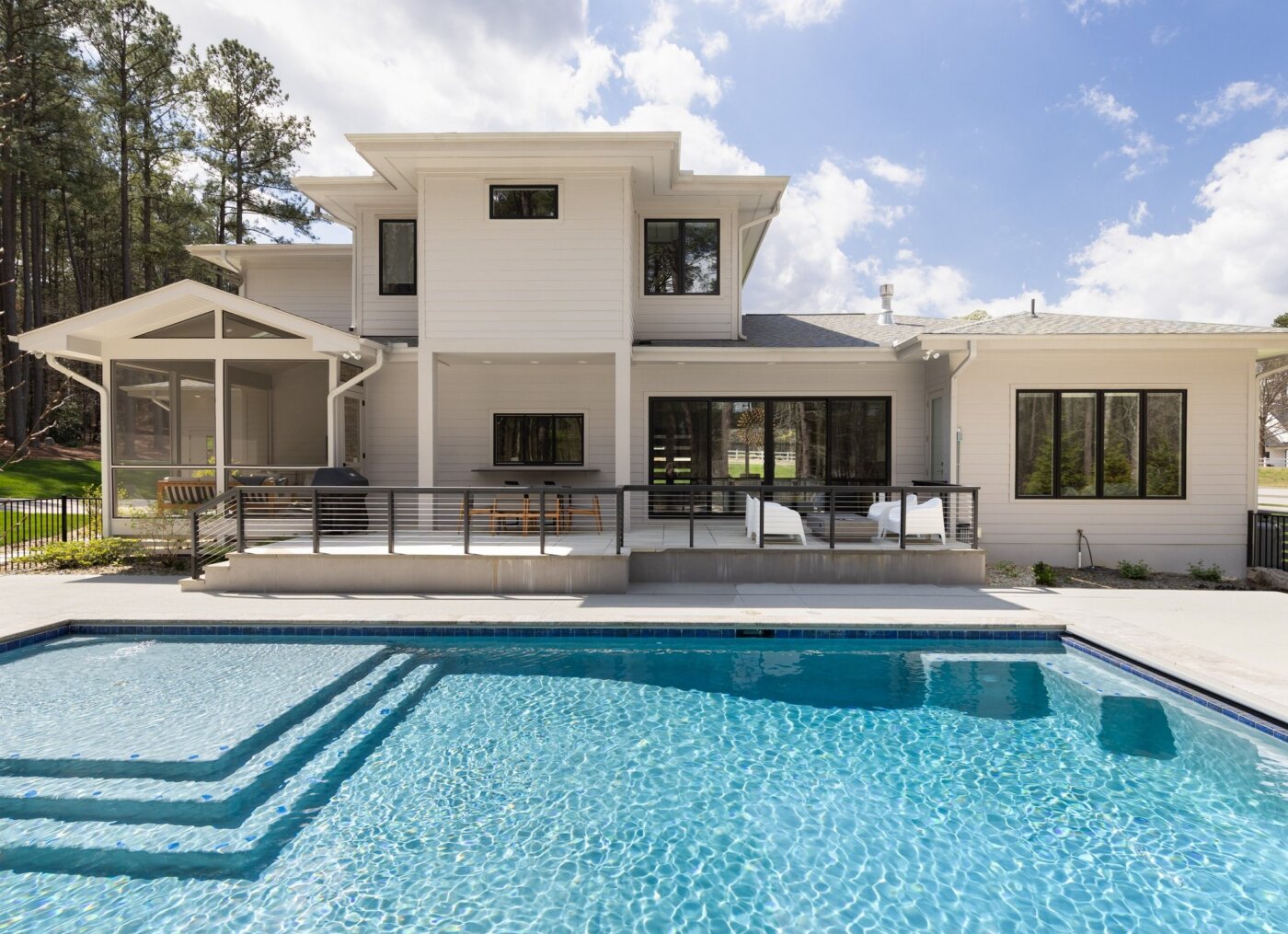 Modern two-story white house with large windows and a covered patio, overlooking a rectangular swimming pool with clear blue water. Surrounded by trees and a bright, sunny sky with a few clouds.
