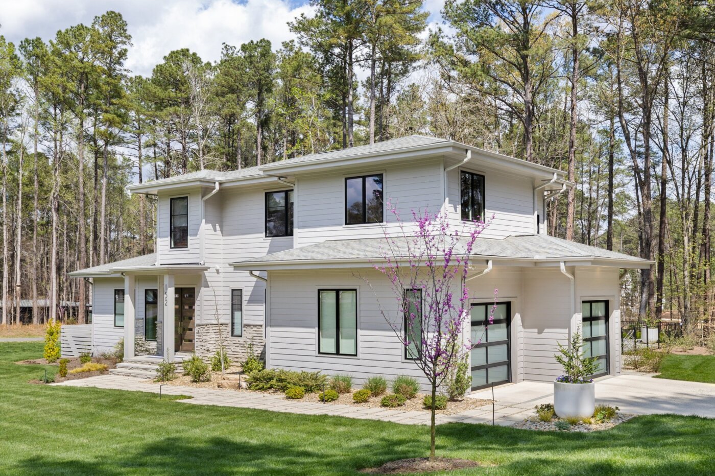 Modern two-story house with large windows, light gray siding, stone accents, and a landscaped yard, surrounded by tall trees under a partly cloudy sky.