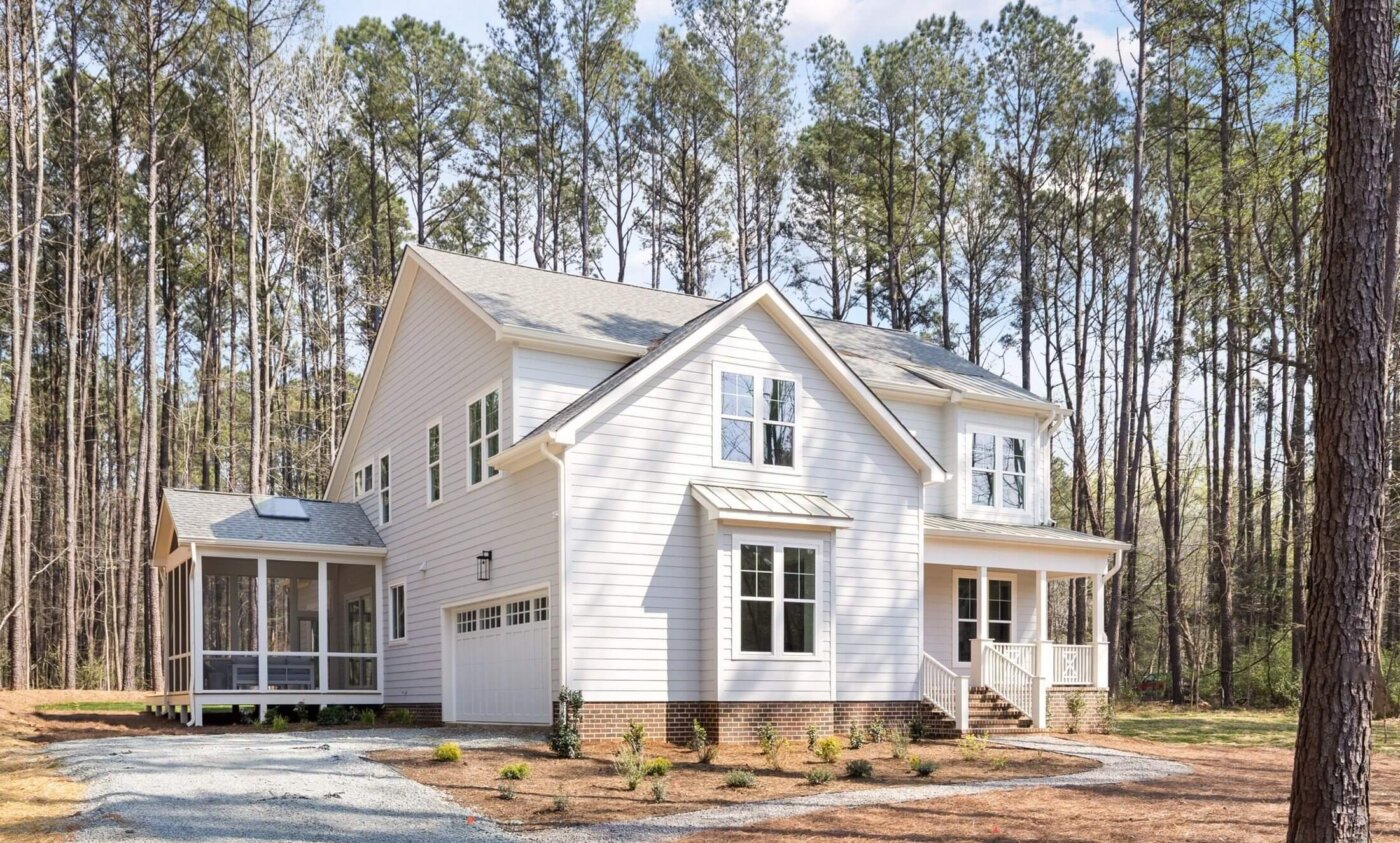 Two-story white house with a covered front porch, attached garage, and screened-in side patio, surrounded by tall pine trees and a gravel driveway.
