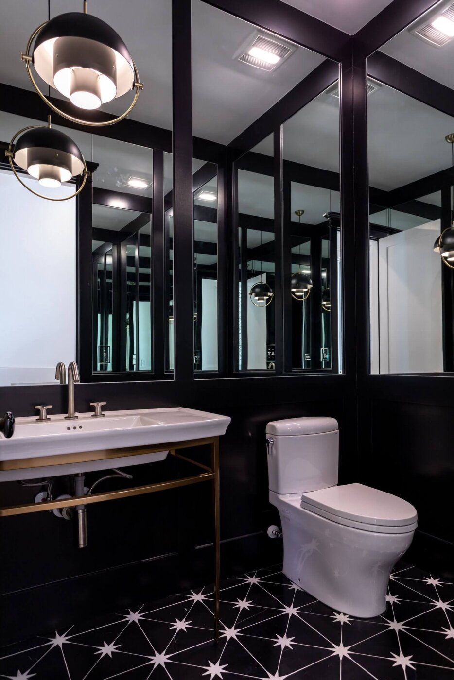 Modern bathroom with black walls, white sink, and toilet. Large mirrors reflect pendant lights and geometric black-and-white tile floor with starburst designs. The space appears sleek and sophisticated.