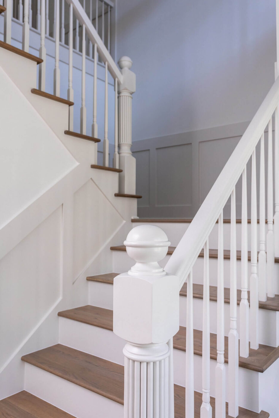 A white staircase with wooden steps, white spindles, and a decorative newel post, set against a light gray paneled wall in a modern interior.
