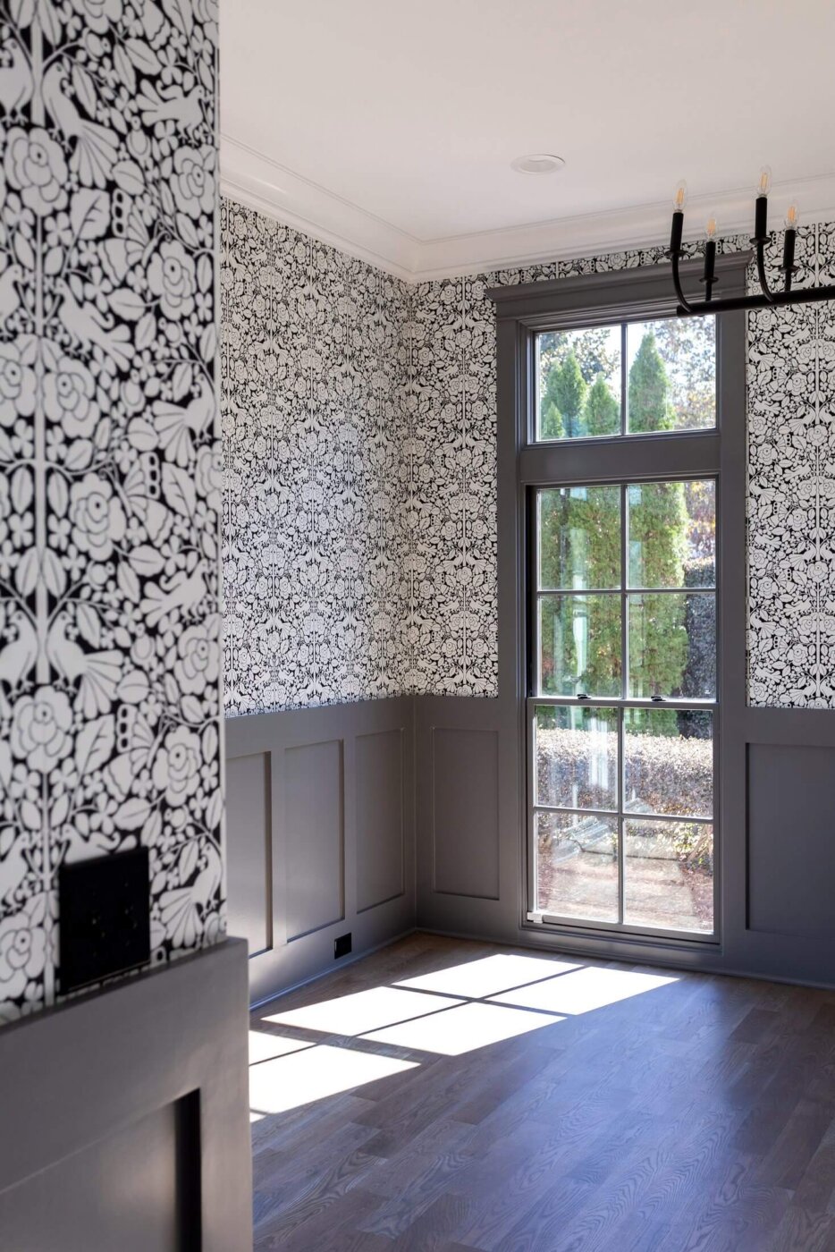 A bright room with black and white floral wallpaper, gray wainscoting, large window letting in sunlight, and a modern black chandelier. Trees are visible outside through the window.