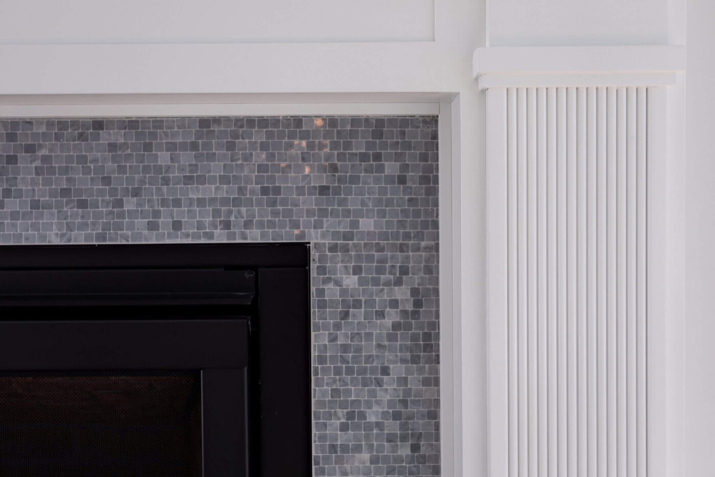 Close-up of a fireplace with small gray mosaic tiles, a black frame, and white decorative molding with vertical grooves on the right side.