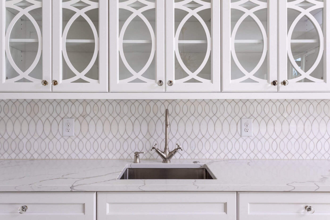 Modern white kitchen with a marble countertop, stainless steel sink, geometric tile backsplash, and glass-front cabinets with circular patterns.