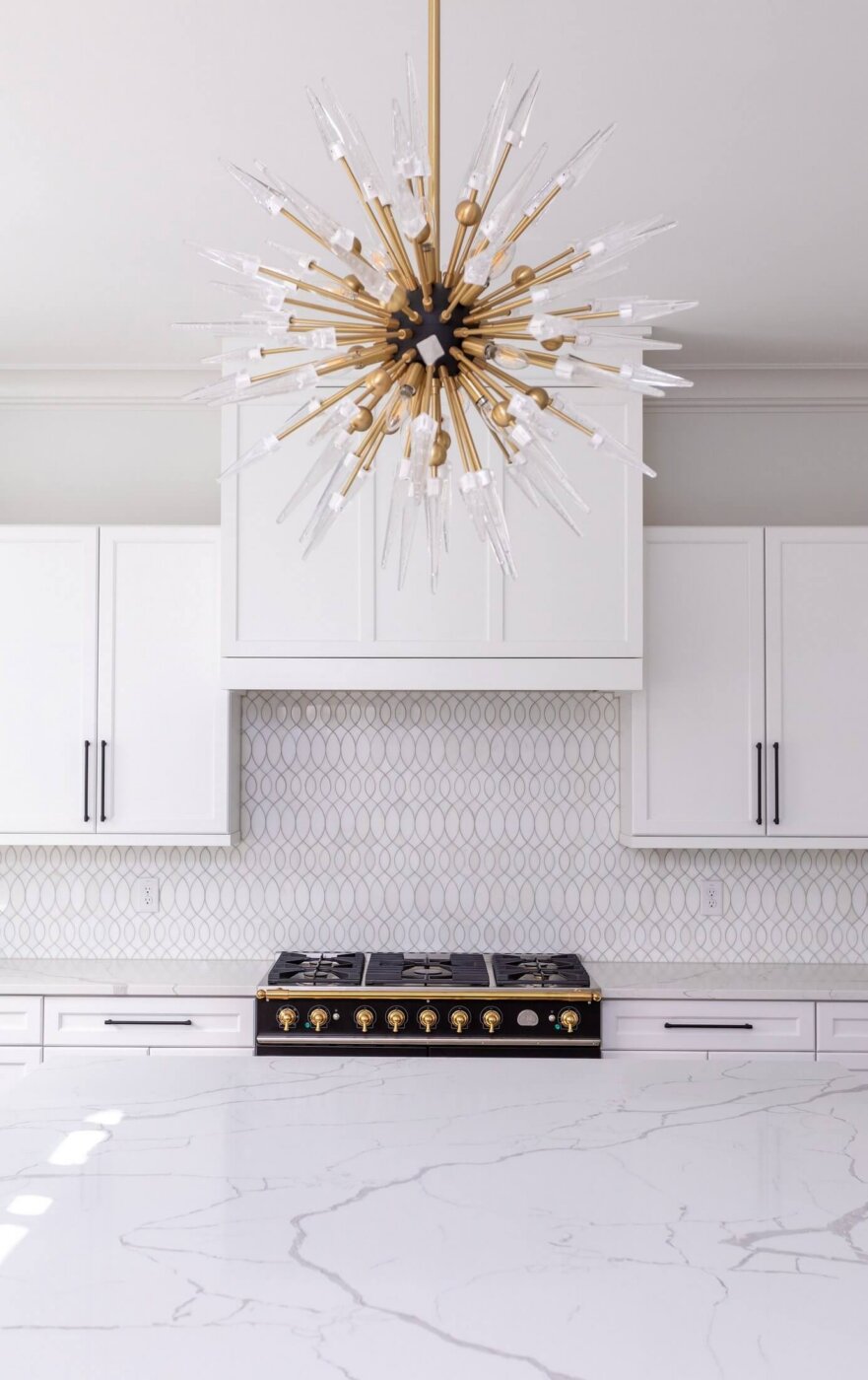 Modern kitchen with a large marble island, white cabinets, a geometric tile backsplash, a black and gold stove, and a striking gold starburst chandelier hanging above.