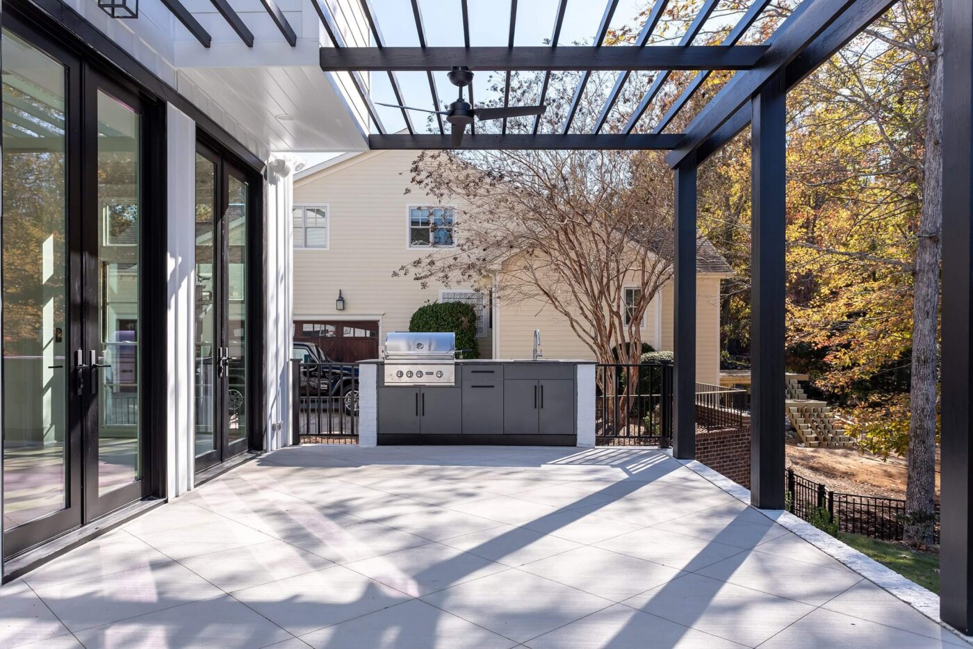 Modern patio with a pergola, outdoor kitchen with a grill, and large sliding glass doors. Trees and a light-colored house are visible in the background on a sunny day.