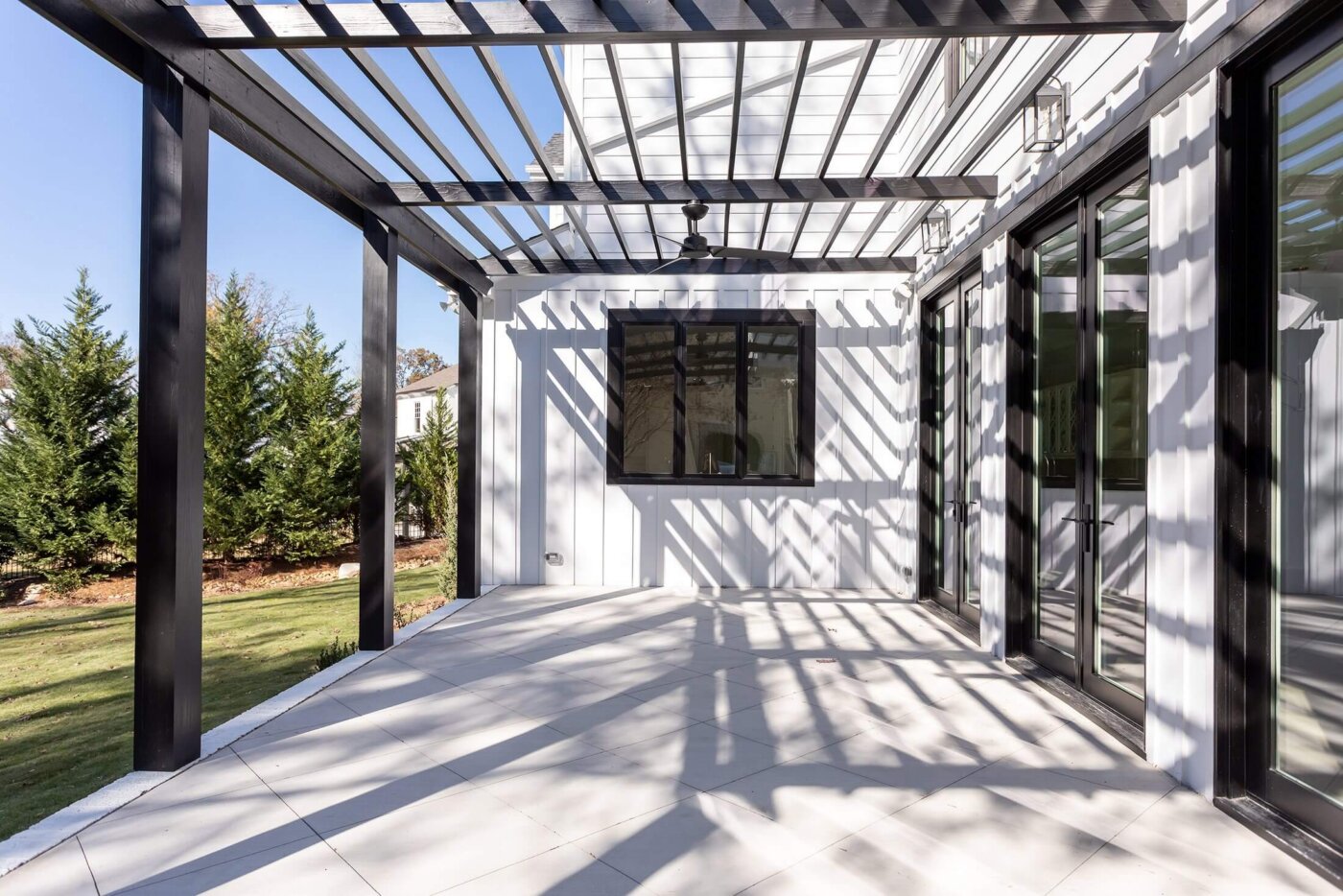 Modern outdoor patio with black pergola casting striped shadows on white tile floor, next to a white house with large windows and glass doors. Green grass and trees visible in the background.