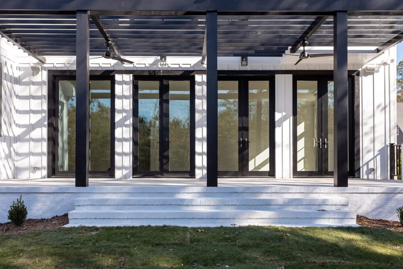 Modern house exterior with white walls, large glass doors framed in black, and a pergola casting striped shadows on the porch steps.