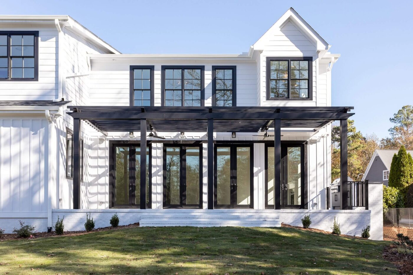 Modern white house with black window frames and a glass-enclosed sunroom, featuring a black pergola and minimal landscaping on a sunny day.