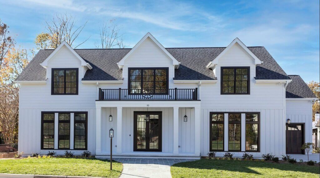 A modern two-story white house with black window frames, a black front door, and a covered entryway with columns, set against a blue sky and surrounded by a green lawn and trees.