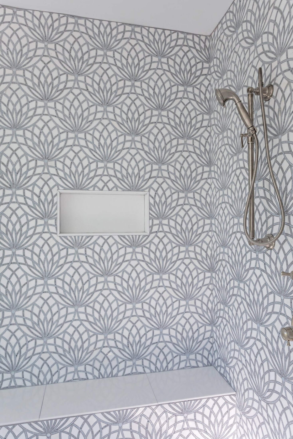 A modern shower with gray and white geometric patterned tiles, a built-in bench, a recessed shelf, and a silver handheld showerhead attached to the wall.
