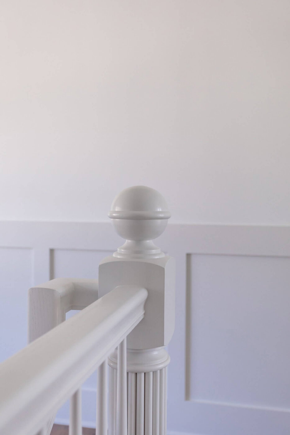 White stair railing with a decorative newel post and a round top, set against a white wall with rectangular molding panels. The overall scene is minimalistic and bright.