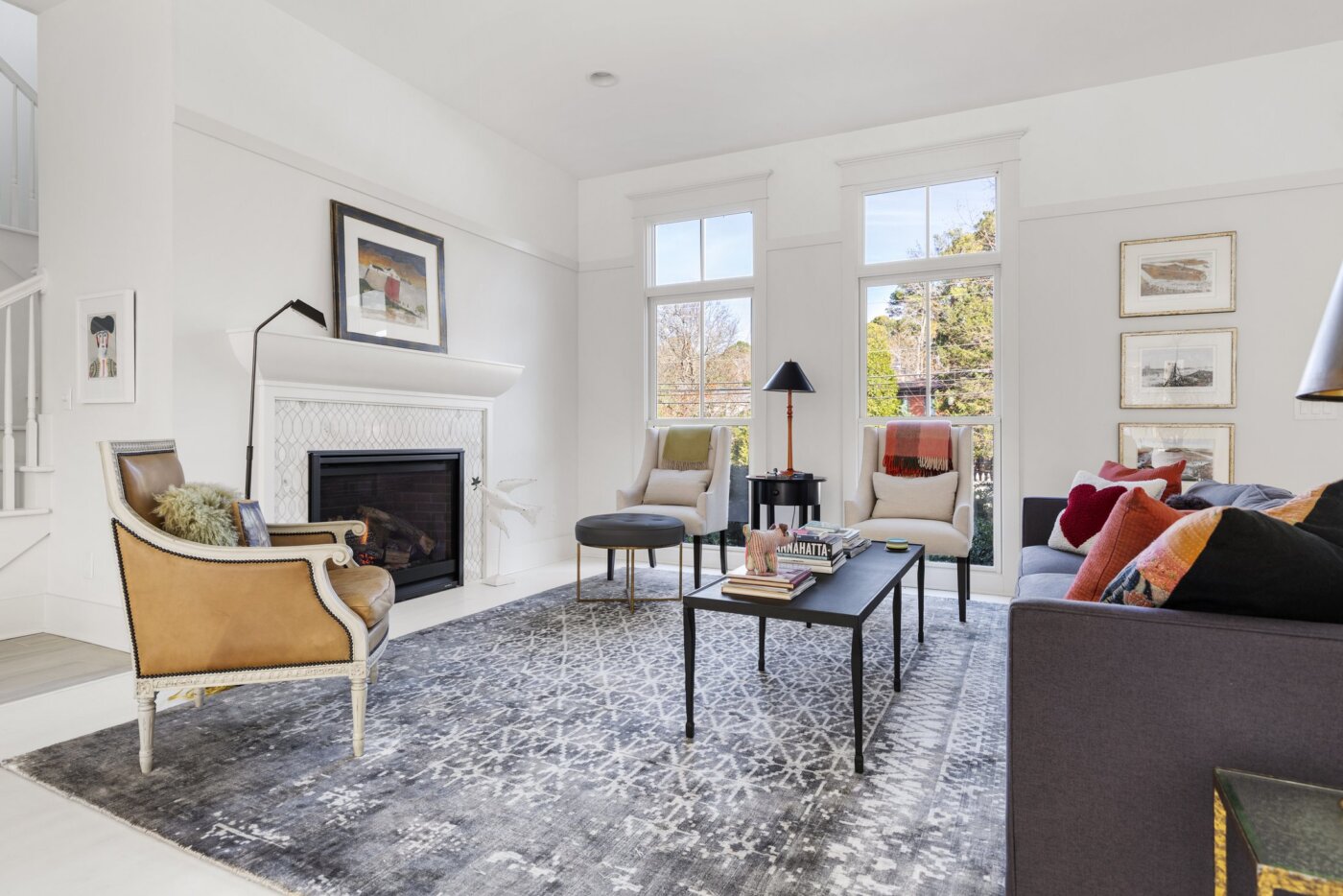 Bright living room with large windows, modern furniture, a patterned rug, fireplace, and artwork on the walls. Coffee table with books and decor, armchairs, and a sofa with colorful pillows create a cozy, inviting space.