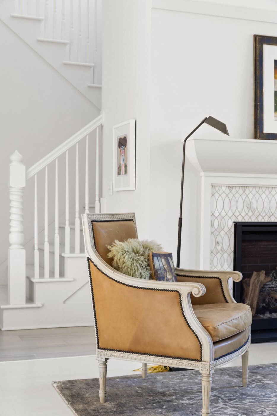 A tan leather armchair with a pillow sits by a fireplace and a tall lamp. Stairs and framed artwork are visible in the bright, white room with light wood floors.