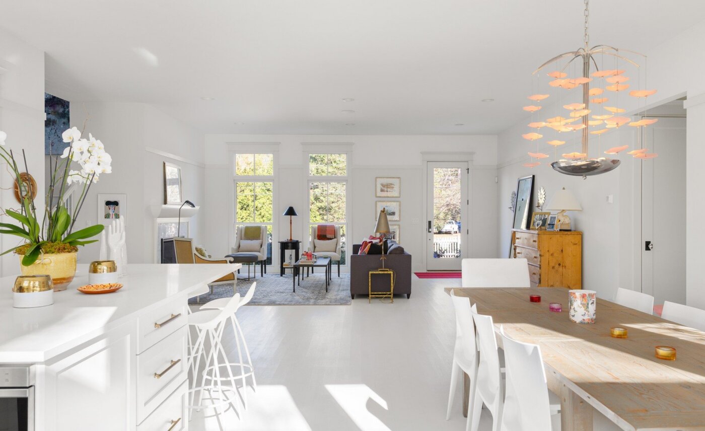 Bright, modern open-concept living and dining area with white walls, contemporary furniture, a wooden dining table, decorative lighting, large windows, and plants.