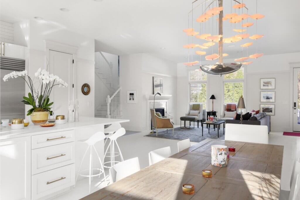 Bright modern open-plan kitchen and living area with white decor, island seating, a wood dining table, orchid plant, and unique orange light fixture. Sunlight streams through large windows creating a cozy, inviting atmosphere.