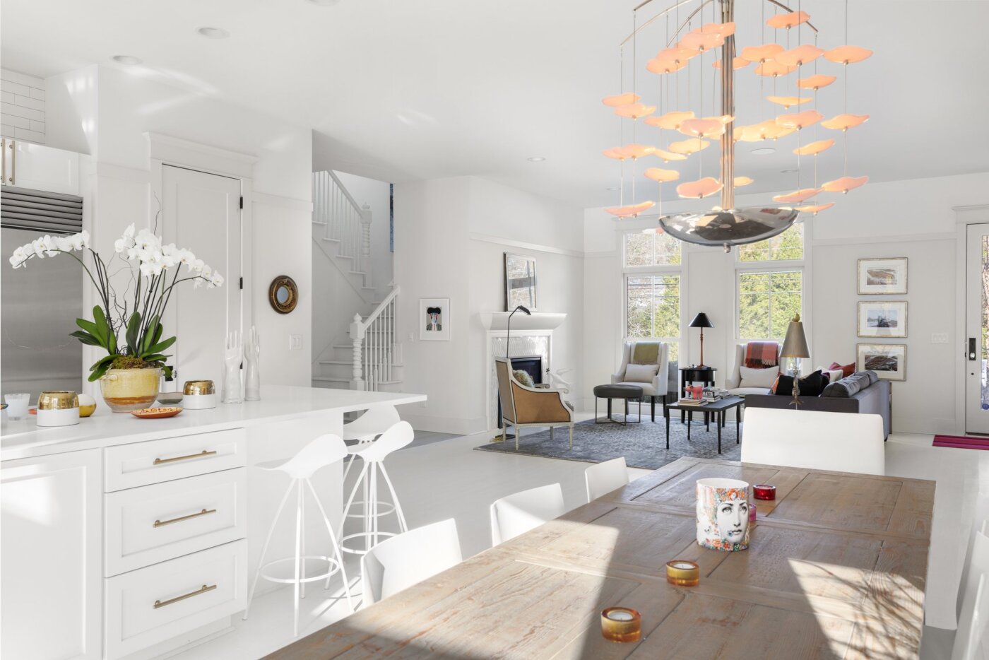 Bright modern open-plan kitchen and living area with white decor, island seating, a wood dining table, orchid plant, and unique orange light fixture. Sunlight streams through large windows creating a cozy, inviting atmosphere.
