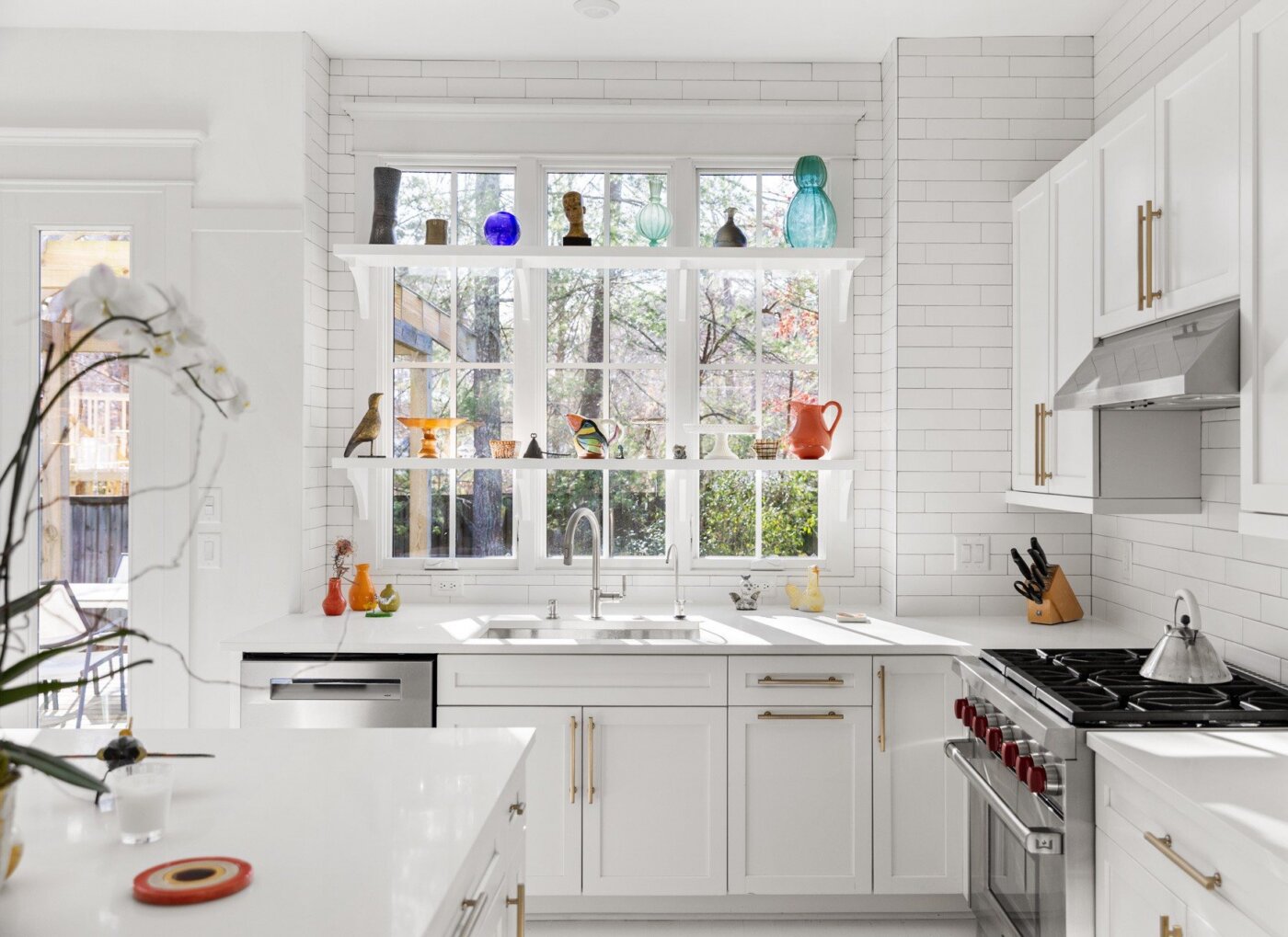 Bright, modern kitchen with white cabinets, tile backsplash, large windows, and open shelves displaying colorful glassware and ceramics. Stainless steel appliances and plants complete the clean, inviting space.