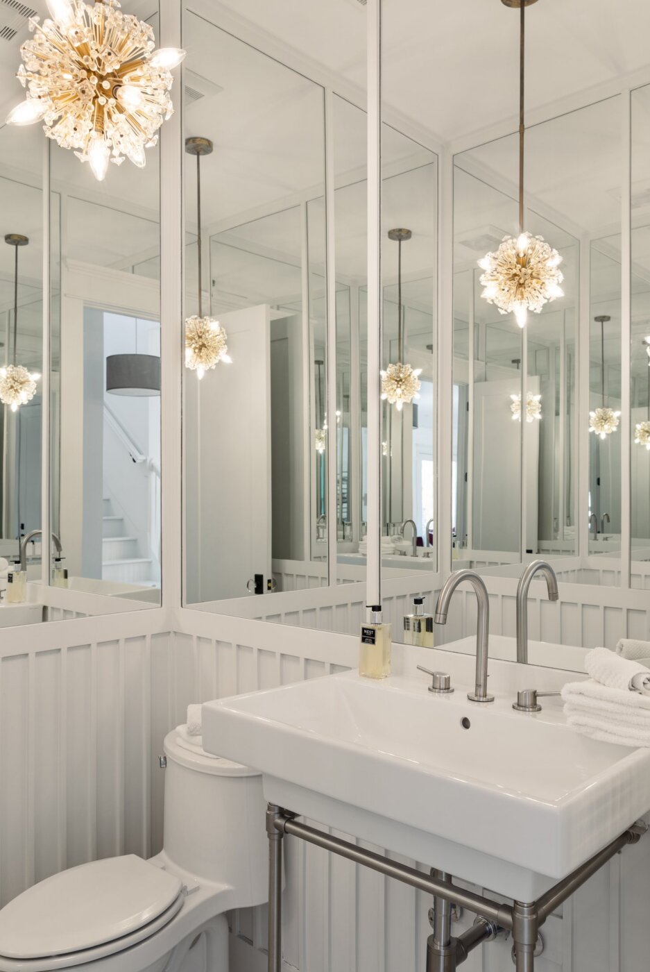 Modern white bathroom with mirrored walls, a rectangular sink, sleek faucet, toilet, towel, soap dispenser, and decorative light fixture with starburst design hanging from the ceiling.