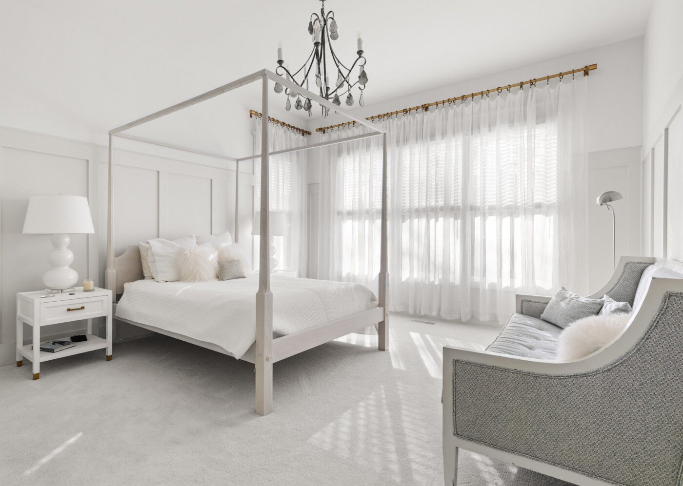 A bright, airy bedroom with a four-poster bed, white bedding, chandelier, nightstand with lamp, elegant gray settee, and sheer curtains letting in natural light.