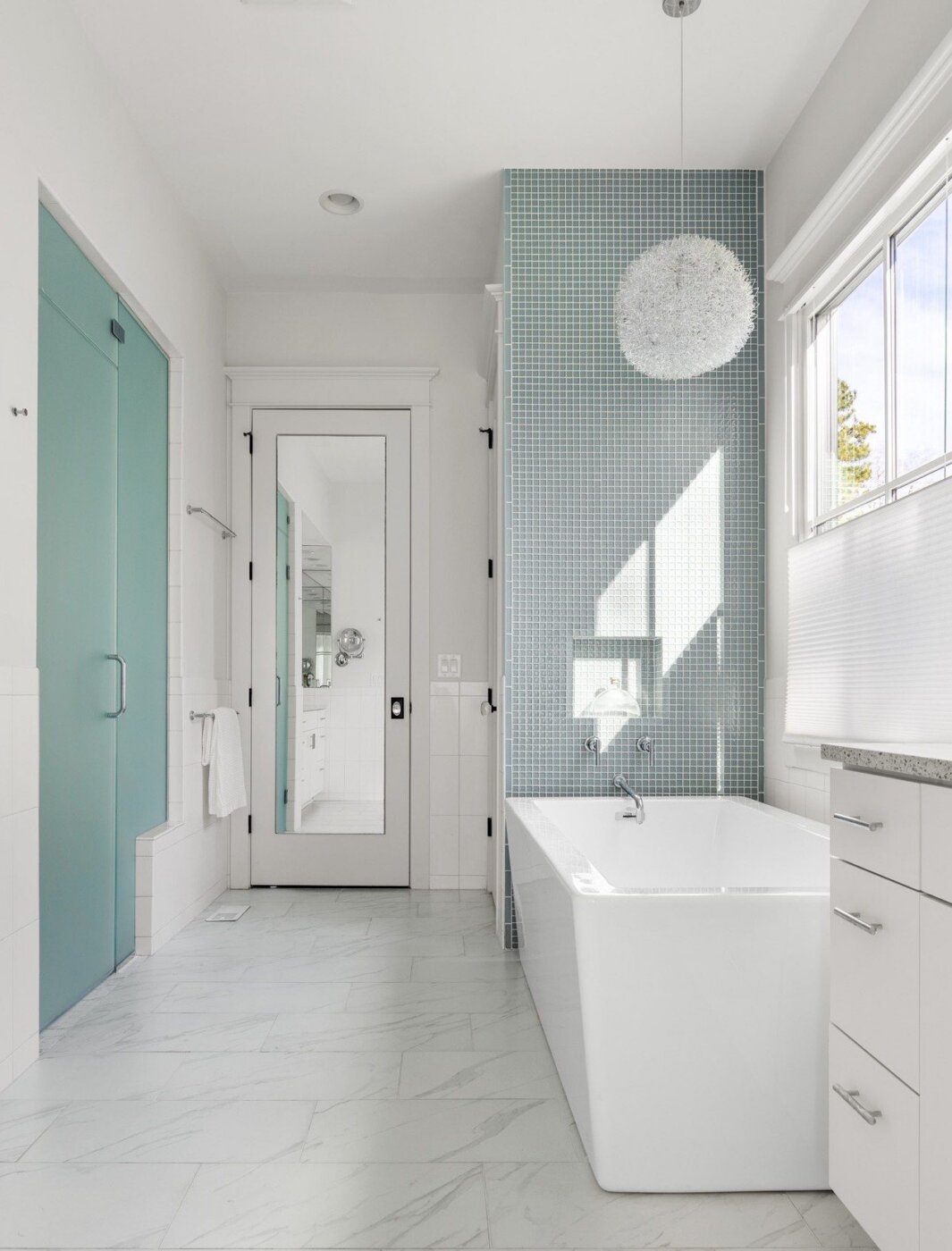 Modern bathroom with white walls and floor, a large bathtub, blue-tiled accent wall, frosted glass shower door, white vanity, large window, and a spherical pendant light above the tub.