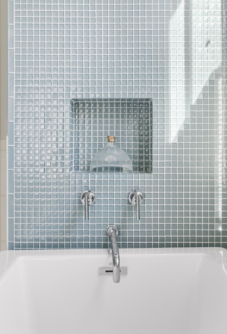 A white bathtub with a chrome faucet and handles is set against a wall of small, light blue tiles. There is a recessed shelf above the tub holding a glass container. Sunlight streams in from the right.