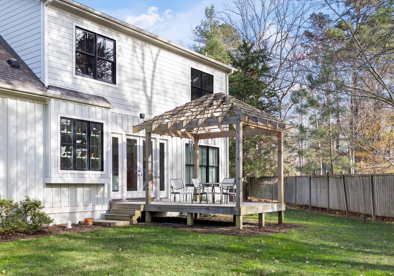 A wooden pergola with patio furniture sits on a raised deck attached to a white house, overlooking a grassy backyard bordered by a wooden fence and trees.