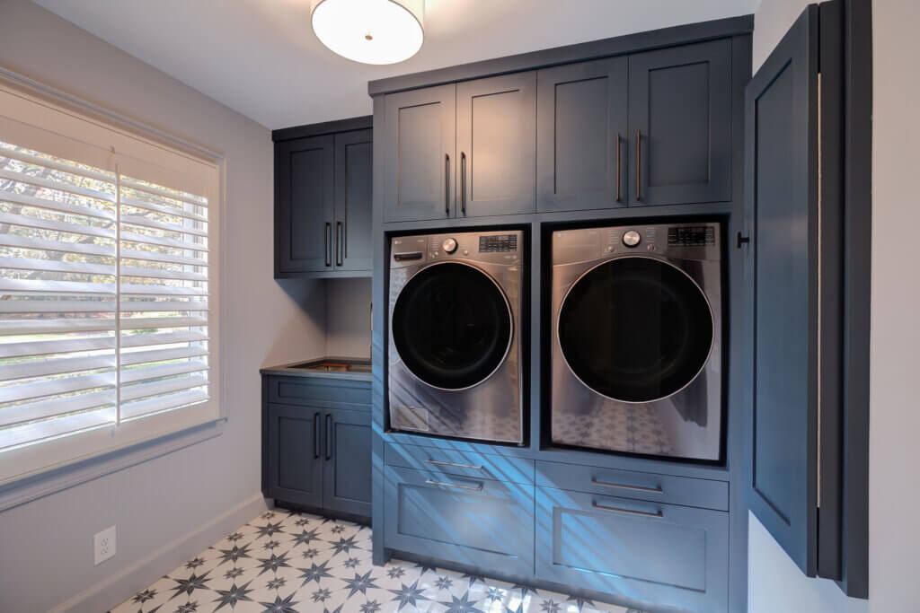 Modern laundry room with blue cabinets, a front-load washer and dryer stacked side by side, patterned tile floor, and a window with white shutters letting in natural light.